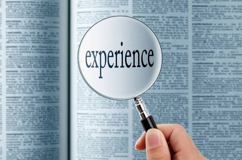This is an image of an open dictionary slightly out of focus taking up the entire image. Above the dictionary is a hand holding a magnifying glass with the word, “experience” magnified.