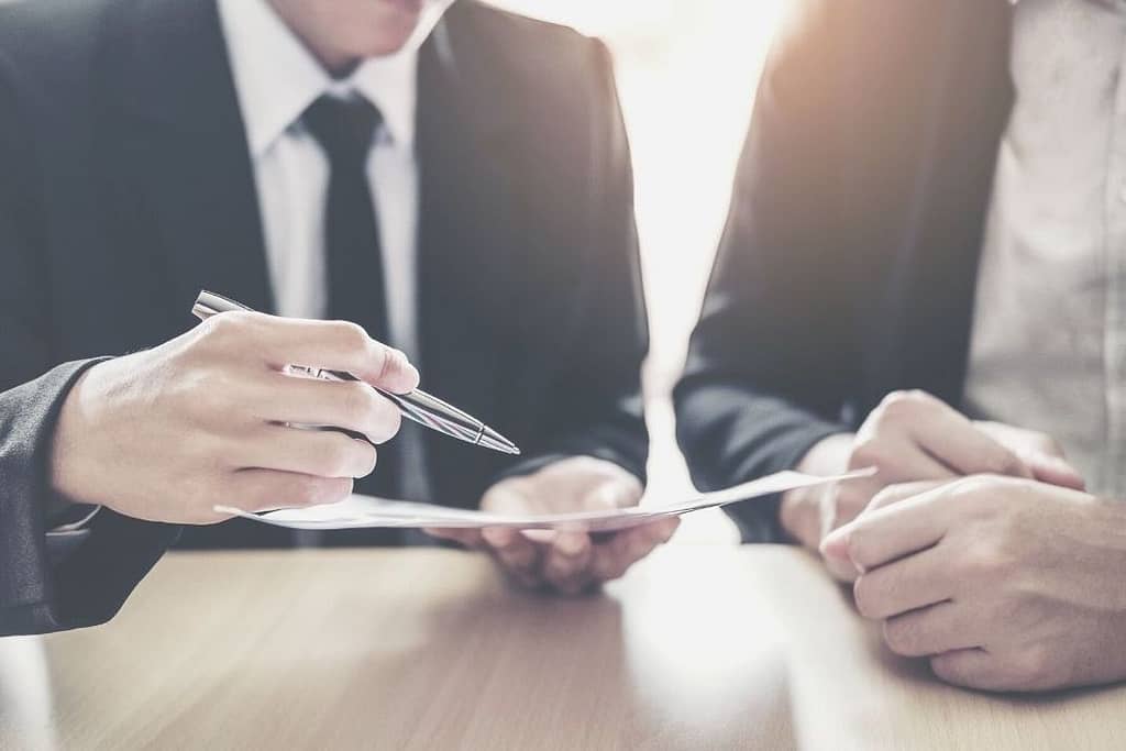 Closeup image of two men sitting at a wooden desk wearing suits and holding nice pens. They are looking over a contract. This image is used for a blog titled "When Hiring a Security Firm, Carefully Read the Security Contract"