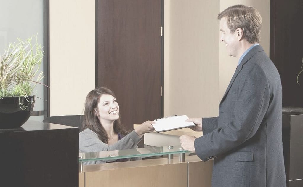 This is an image of a man wearing a business suit checking in at a front desk at a company’s office.This mage is used for the BPS Security article titled, “How Visitor Policies Can Increase Security Risk.”