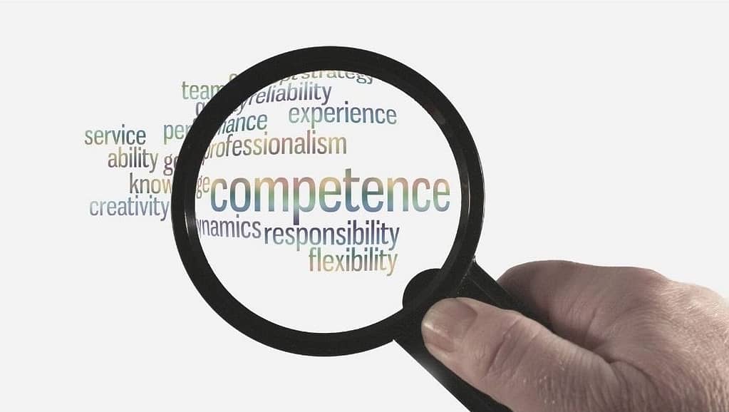 This is an image of a hand holding a magnifying glass scanning over a word cloud that has the word “competence” along with other similar words. This image is used in the BPS Security article titled, “Why Making Price the Determining Factor Leads to Fatalities”.