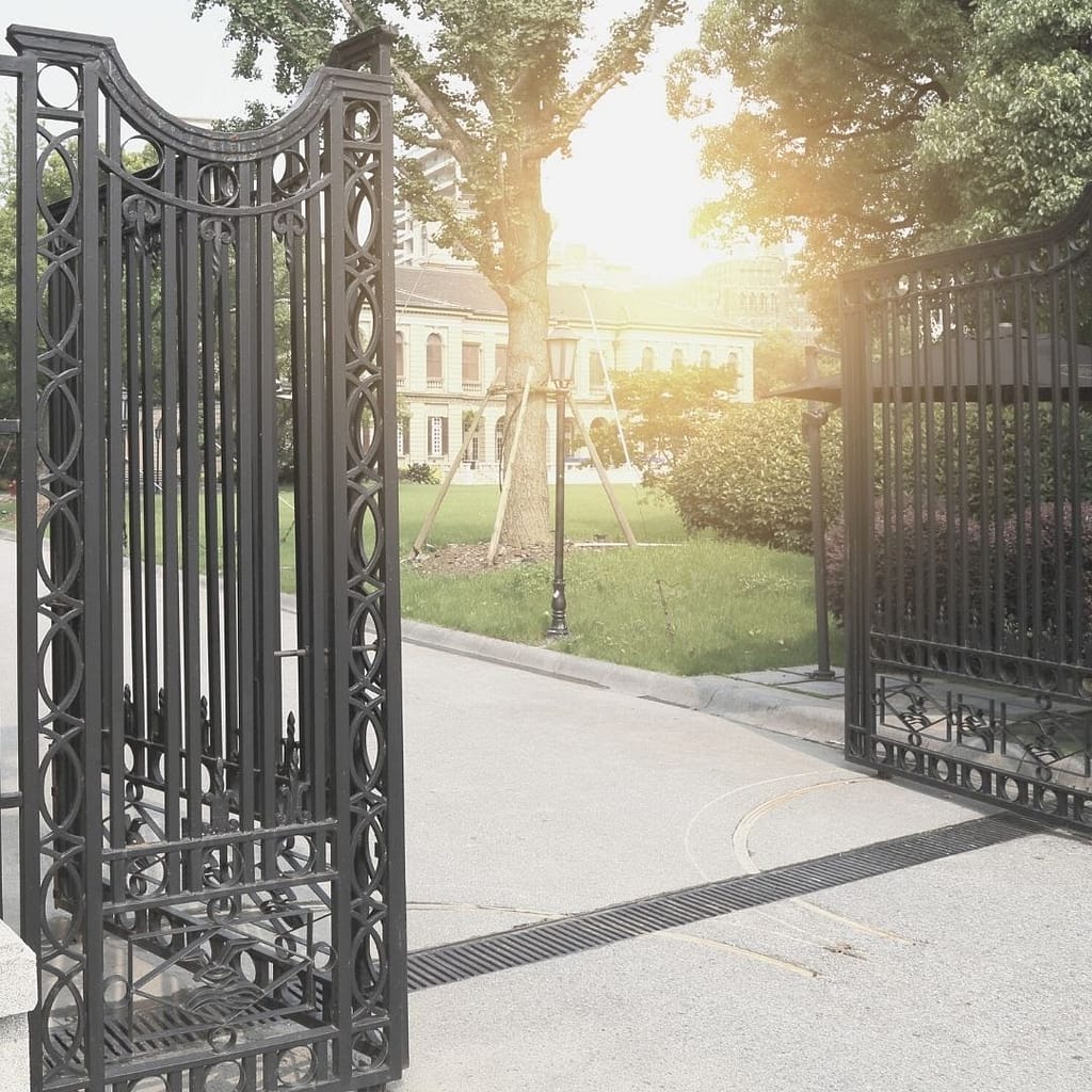 Image of a rod iron gate that is open. You see a tree behind the gate with the sun peaking around the tree. This image is use for the BPS Security Blog How Environmental Design Can Reduce Crime
