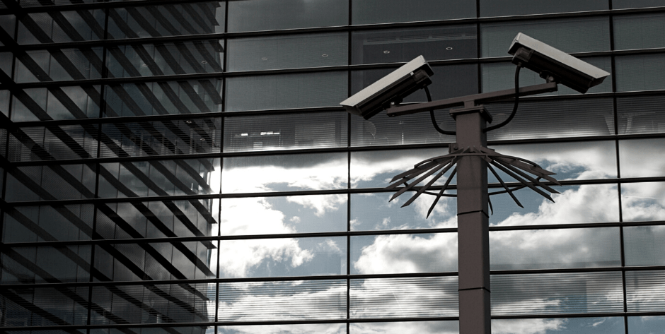 Image of two security cameras on top of a pole in front of a large mirrored building with clouds in the reflection of the mirrors