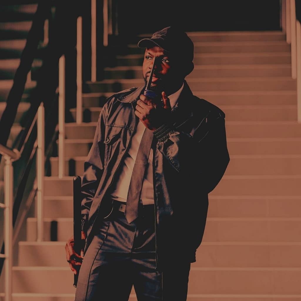 This is an image of a security guard standing at the base of a flight of stairs in a building holding a walkie talkie near his mouth. This image is used in the BPS Security Blog titled, “9 Security Firm Stories that Will Give You CHILLS!”.