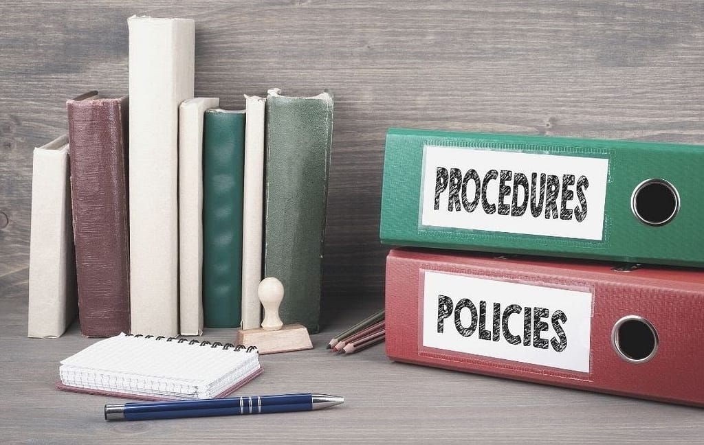 Photo of a desk with books stacked up agains the wall and two large binders reading, "procedures" and "policies. This image is used in the BPS Security Blog titled, "The Importance of Security Culture."