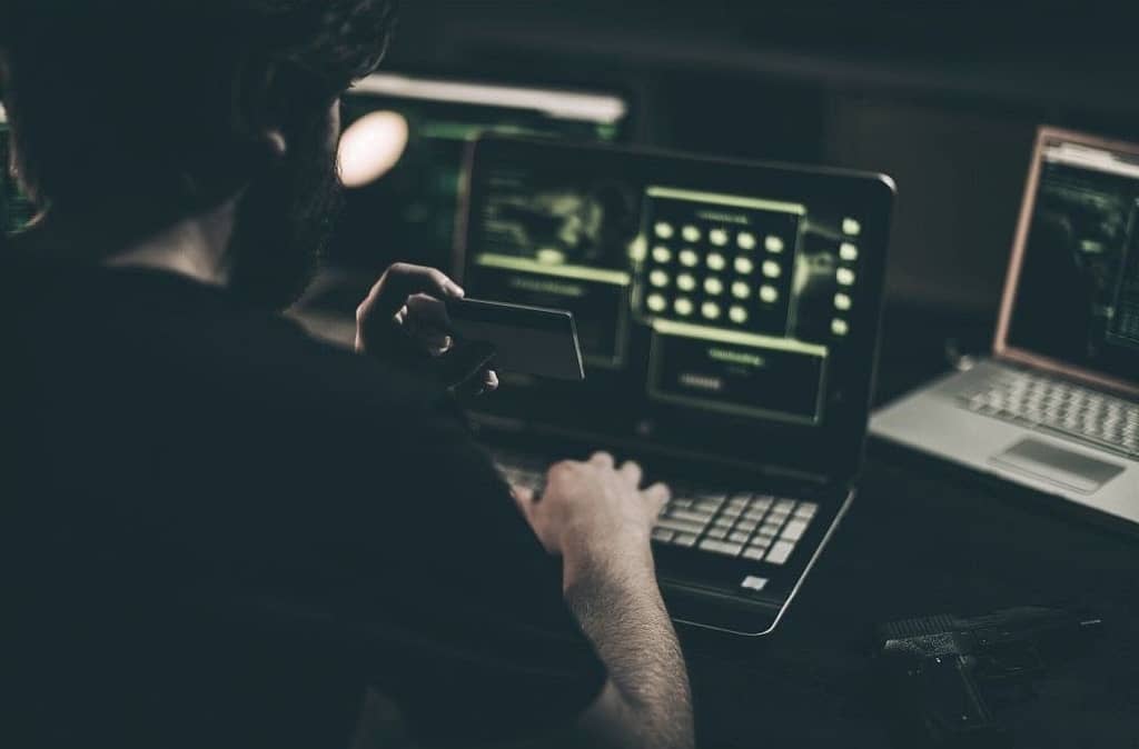 This image showcases a company’s employee sketchily (in dim light) hacking into a company computer and holding what is likely not his credit card.. This image is included in the San Antonio-based, BPS Security’s article titled, “How Company Culture Affects Security”.